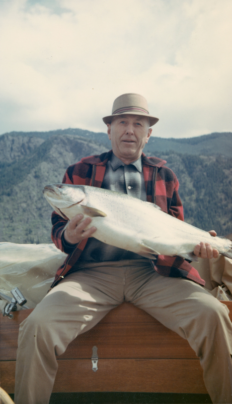 A man wearing a plaid jacket and a hat is sitting on a wooden surface, holding a large fish. There are mountains visible in the background.