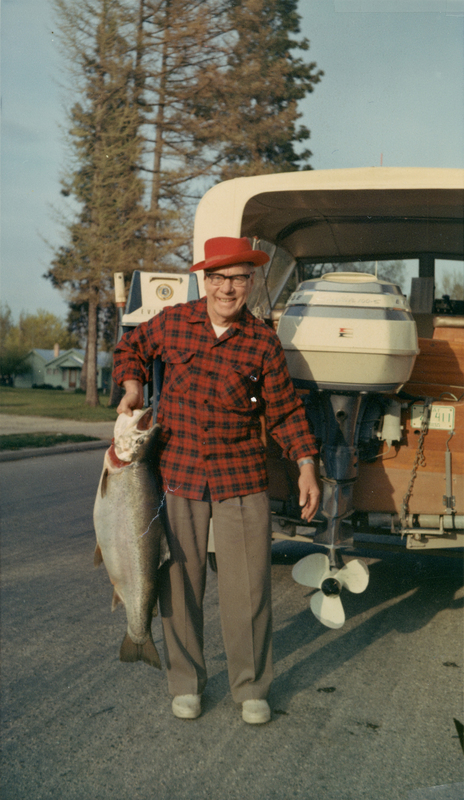 A person wearing a red hat and plaid shirt is standing on a street holding a large fish. Behind them is a vehicle with a boat and an outboard motor. Trees and houses are visible in the background.