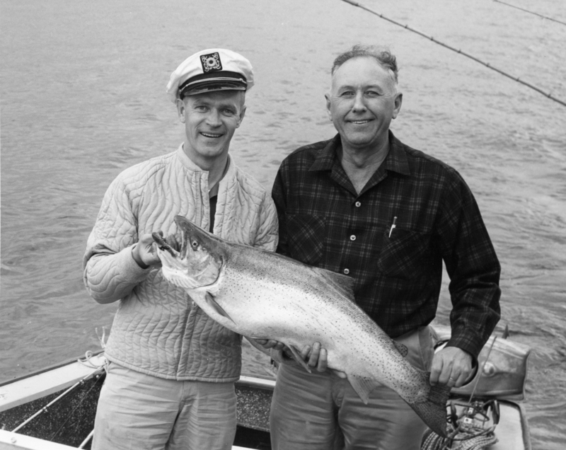 Two men standing on a boat holding a large fish. One man is wearing a quilted jacket and captain's hat, and the other is wearing a plaid shirt. The water is visible in the background. Label on cap reads "USS".