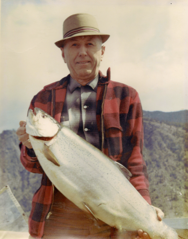A person standing outdoors, wearing a checkered jacket and a hat, holding a large fish. Mountains can be seen in the background.