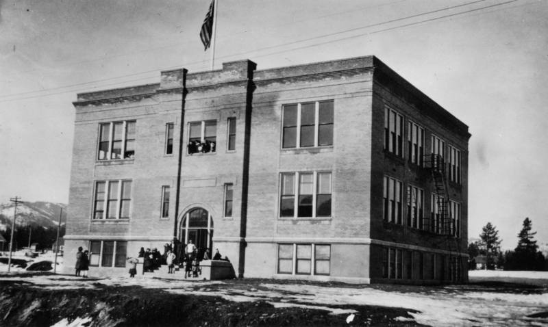 A large brick building with multiple windows and a flagpole on the roof. A group of people, mostly children, are gathered near the entrance of the building. Snow covers the ground, and there are trees and hills in the background.