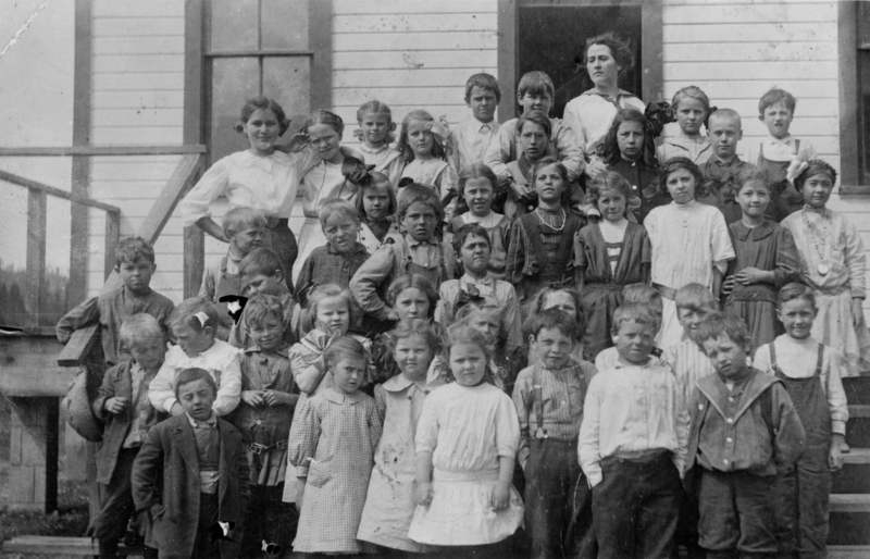 A group of children standing in rows on the steps of a wooden building, with two adults behind them. The children are dressed in early 20th century clothes, some with ribbons in their hair and others wearing suspenders or jackets.
