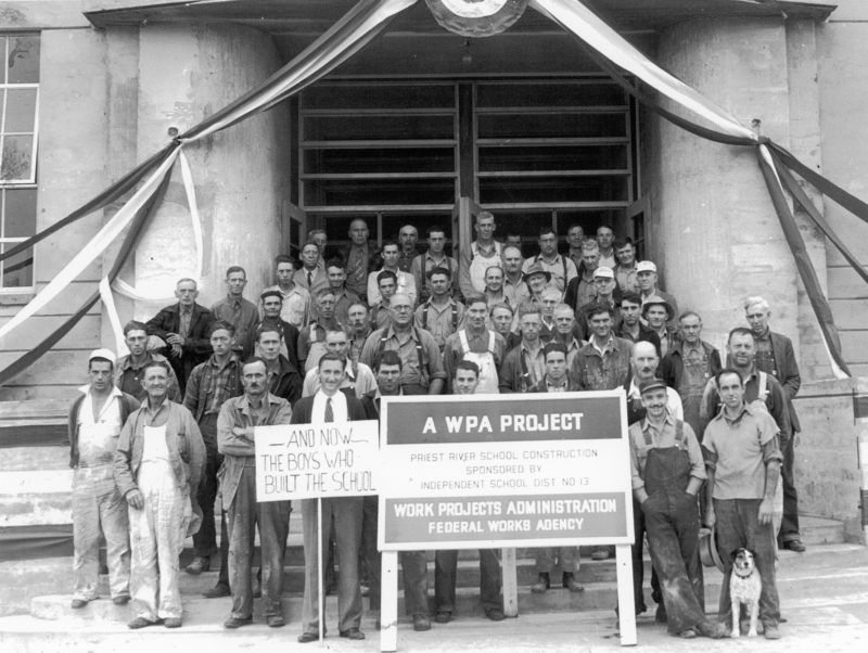 A large group of men standing on the steps of a building with two signs in front of them. "- Sign near the center: "A WPA PROJECT." - Below that: "PRIEST RIVER SCHOOL CONSTRUCTION SPONSORED BY INDEPENDENT SCHOOL DIST NO 13." - At the bottom: "WORK PROJECTS ADMINISTRATION FEDERAL WORKS AGENCY." - Sign to the left: "AND NOW- THE BOYS WHO BUILT THE SCHOOL."