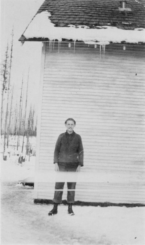 A person stands in front of a building with wooden siding. Snow covers the roof, and there are icicles hanging down. The ground is snowy, and there are trees in the background.