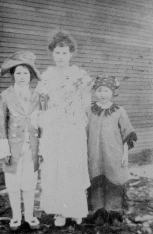 Three children dressed in costumes stand in front of a wooden wall. The child on the left wears a tricorne hat and a coat, the one in the middle wears a light-colored dress, and the child on the right wears a headscarf and a fringed garment.