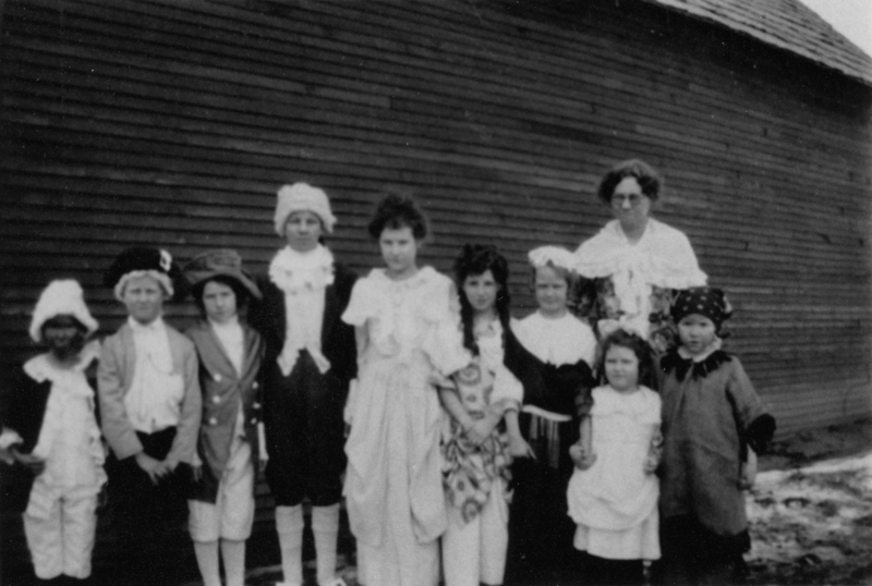 A group of children and an adult wearing costumes, standing in front of a wooden building. The children are dressed in various outfits, including hats and ruffled garments. The adult stands behind them, observing with a calm demeanor.