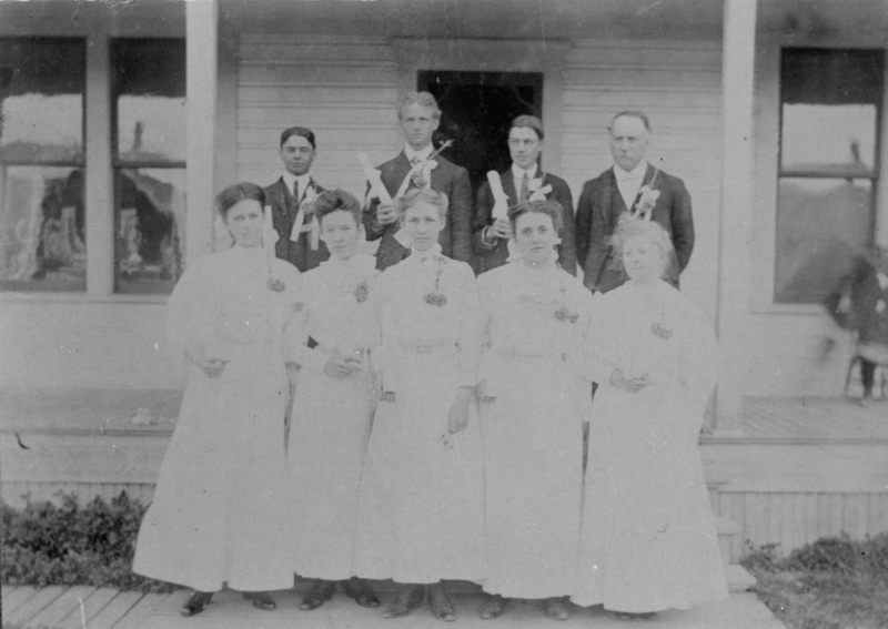 A group of eight people stands on the porch of a wooden building. Four women wearing long dresses with flowers pinned to them are in the front row. Behind them, four men in suits, three of whom hold rolled documents, are positioned.
