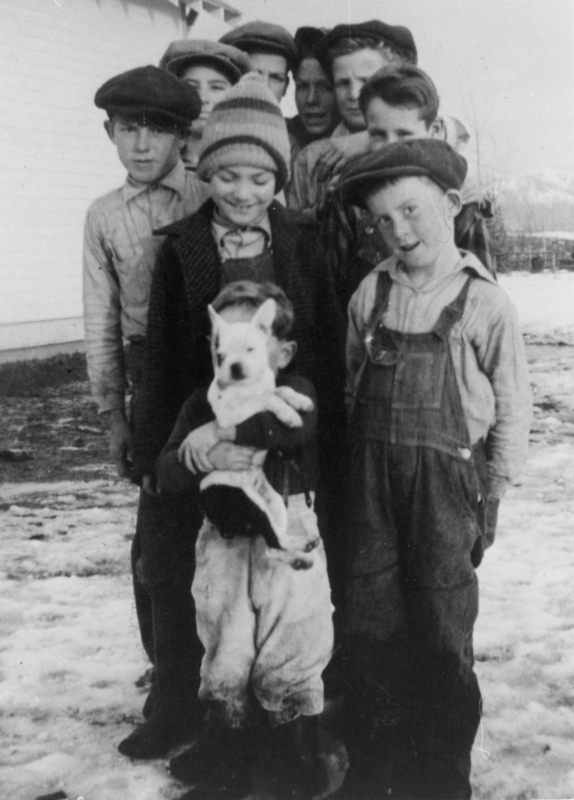 A group of boys standing together, with some wearing hats and overalls. One boy at the front is holding a small dog. The background includes a building and snow on the ground.