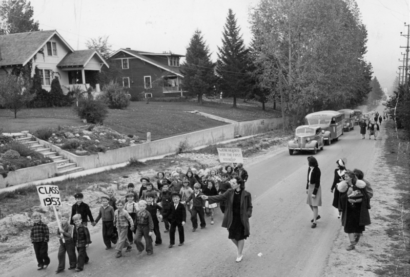 A group of children walking along a street, led by a woman pointing forward. In the background, several school buses are parked along the side of the road. There are houses and trees lining the street. Another adult and two other children are also visible nearby. Sign held by child reads: "CLASS OF 1953" - Sign in the background reads: "WE'RE HEADED FOR THE FIRST ROUNDUP"