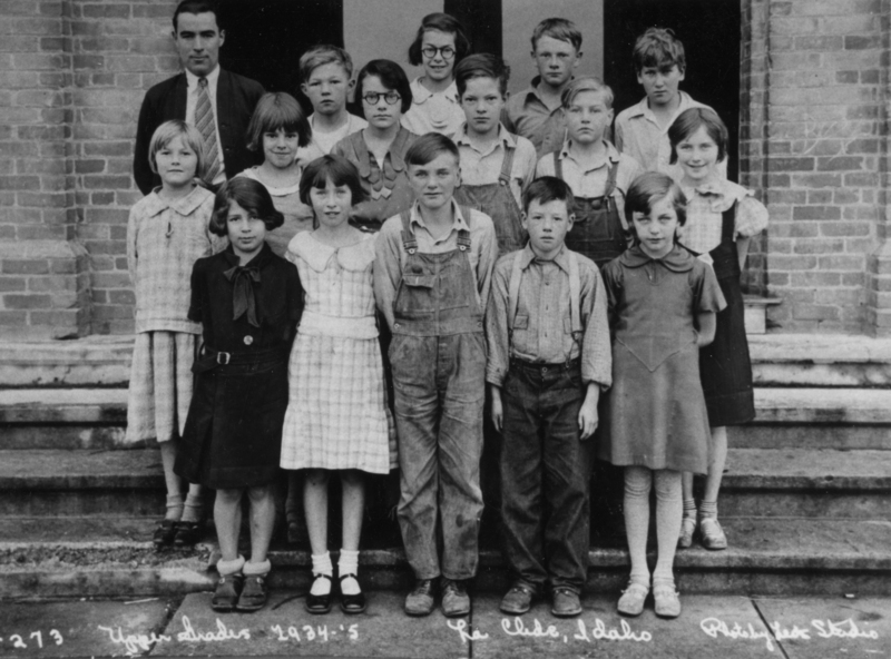 A group of children and an adult stand on the steps in front of a brick building. The children are dressed in various outfits, including dresses and overalls. The adult is wearing a suit and tie.