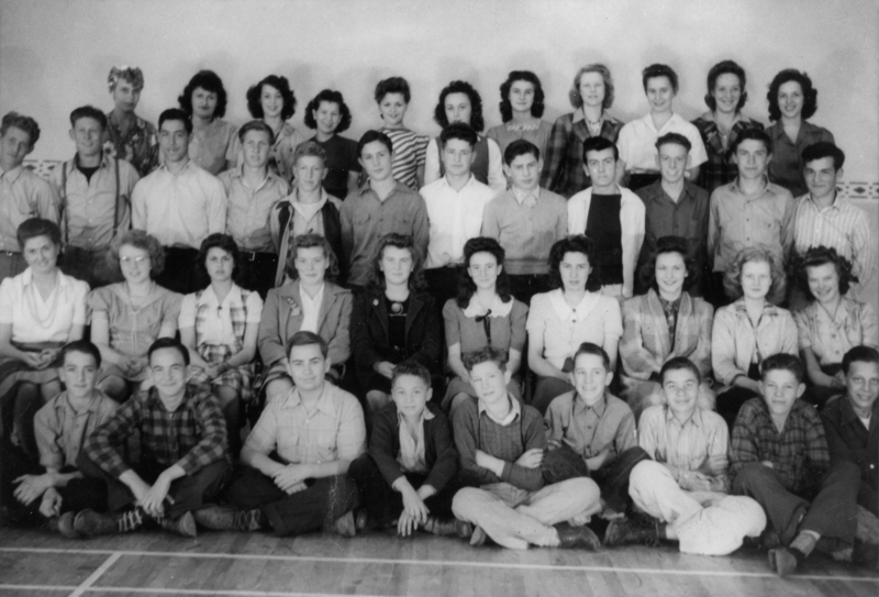 A group of young men and women posing together in several rows. The individuals are dressed in mid-20th century clothing, with some sitting on the floor and others standing behind them. The setting appears to be indoors.