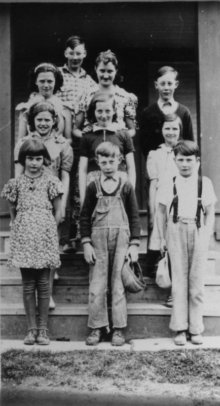 A group of nine children standing in rows on wooden steps. The children are wearing various outfits, including dresses, overalls, and shirts with suspenders. Some of the children are holding items like hats or bags. They are posed in a relaxed manner, with some of the children smiling.