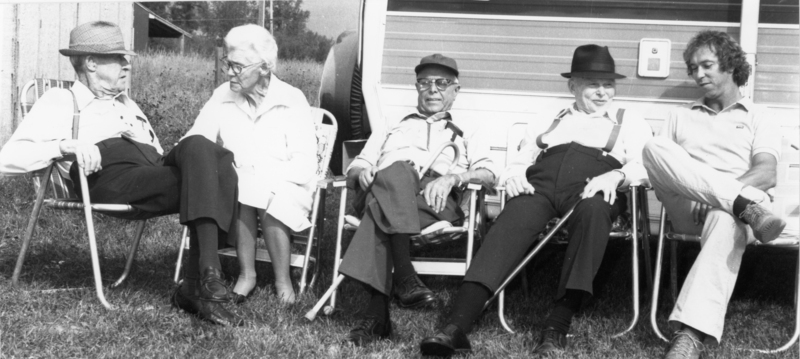 Five people are sitting in lawn chairs outside. They are positioned in a line next to a trailer, with three older men wearing hats and suspenders on one side, and one older woman with glasses and a younger man wearing a polo shirt on the other. The setting appears to be a grassy area with a building and trees in the background.