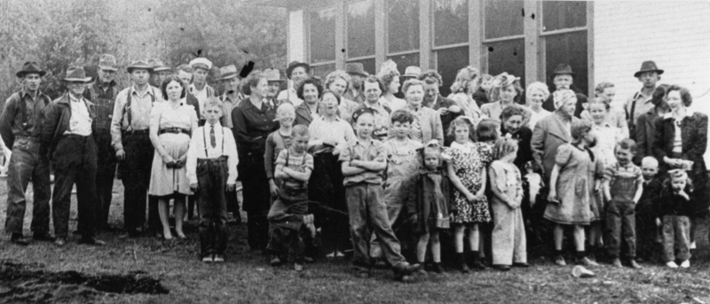 A large group of men, women, and children standing together outdoors in front of a building with windows. Some are wearing hats and there is a mix of casual and formal clothing styles. The children are at the front of the group.