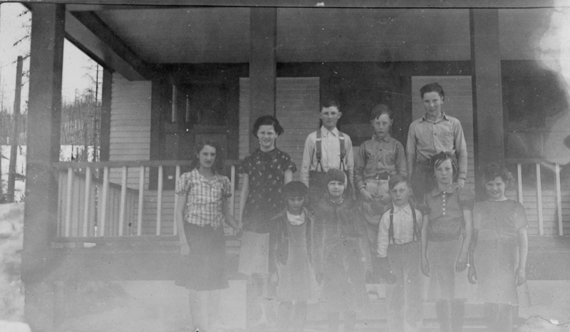A group of ten children standing on the steps of a wooden porch, with a house visible in the background. The children are dressed in various styles of clothing common in the early to mid-20th century.