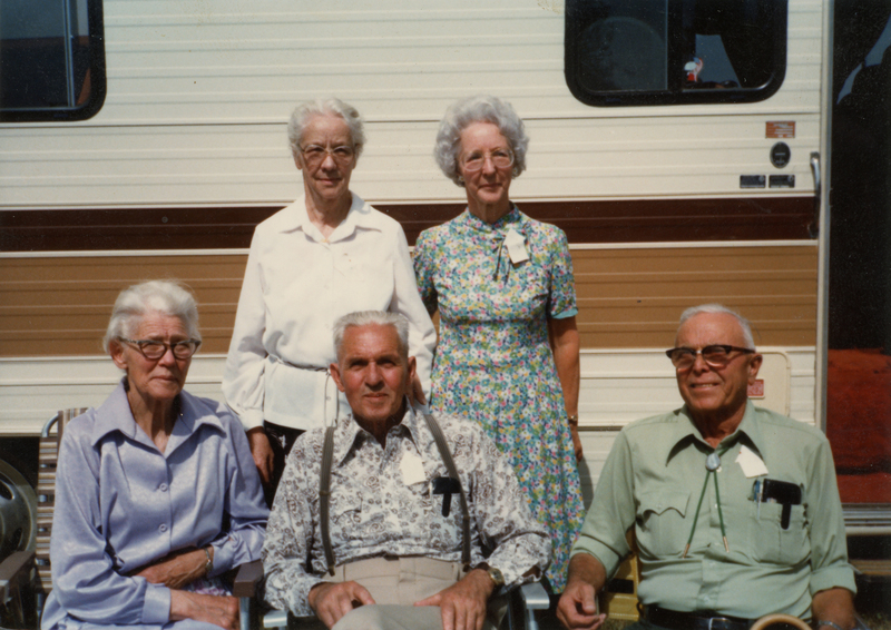 A group of five older adults posing in front of a camper. Two women stand in the back; one is wearing a white blouse, and the other has a floral dress. In the front, a woman in a purple blouse and two men, one in a patterned shirt with suspenders and the other in a light green shirt, are sitting.