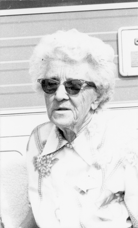 An elderly woman with curly hair is wearing sunglasses and a patterned blouse. She appears to be sitting in front of a textured surface.