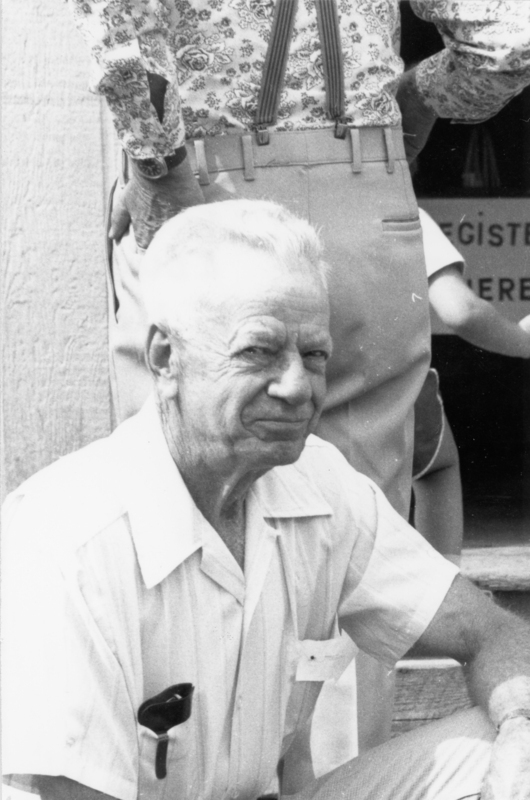 An elderly man with short white hair is sitting and wearing a light-colored collared shirt with sunglasses in the pocket. Behind him, another person is standing, wearing pants with suspenders and a floral-patterned shirt. There is a partial view of a sign in the background.