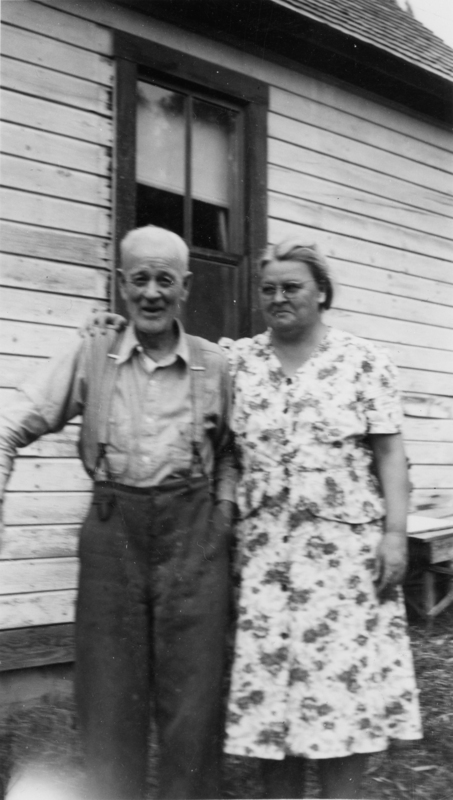 An older man and woman standing in front of a wooden building. The man is wearing suspenders and a button-up shirt, and the woman is in a floral dress, with her arm around his shoulder.
