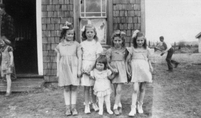 Five young girls stand in front of a small wooden building. The central girl is holding hands with a smaller child. All the girls are wearing dresses with bows in their hair. In the background, there is a man watching from the building's entrance, a young girl beside him, and another young boy playing in the field nearby.