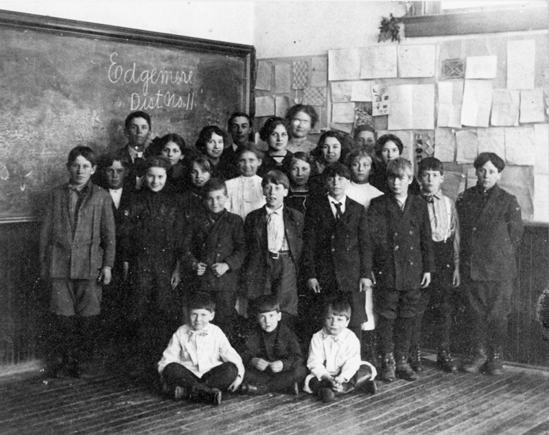 A group of children and a few adults standing in a classroom. Behind them is a blackboard with writing on it and a wall covered with drawings or papers. The floor is wooden.