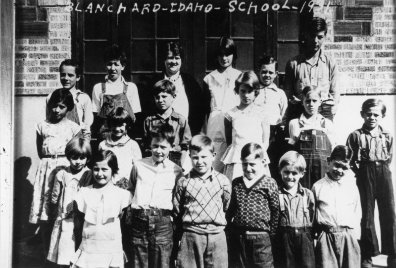 A group of children and one adult are standing in front of a brick building. The children are arranged in rows, with some wearing dresses and others in shirts and overalls. The adult is standing in the middle of the second row. The building has large windows and a patterned brick wall. The text near the top reads "BLANCHARD - IDAHO - SCHOOL - 1921".