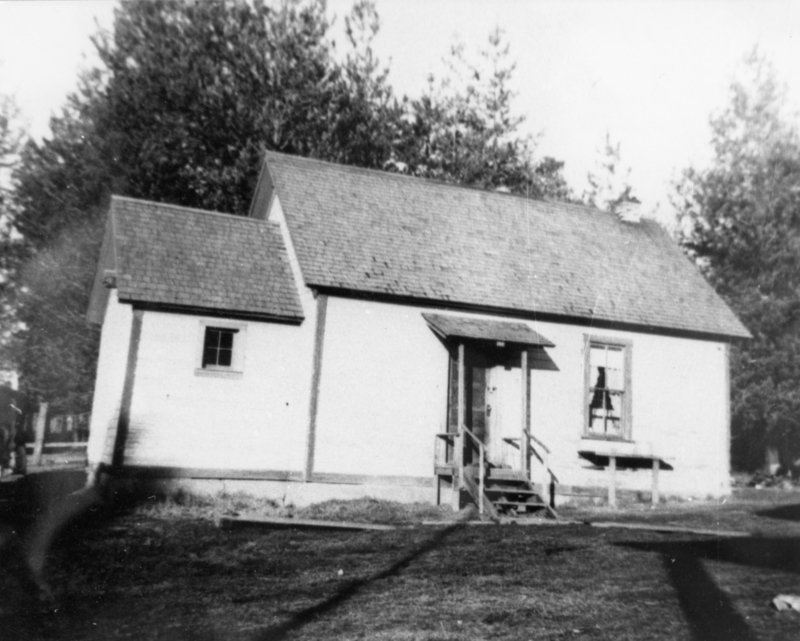 A small wooden building with a peaked roof, front steps, and two visible windows. Trees can be seen in the background.