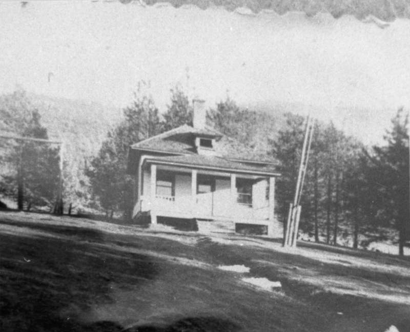 A small house with a porch sits on a slightly sloped grassy area, surrounded by trees.
