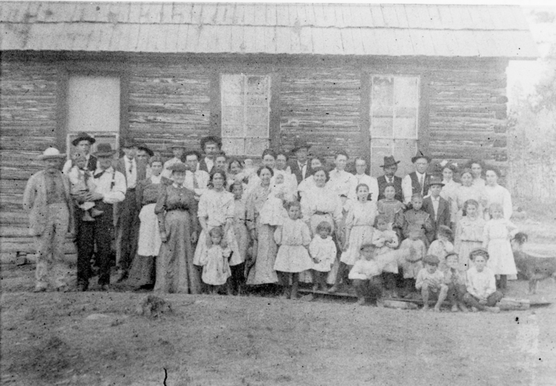 A large group of men, women, and children standing and sitting in front of a log building with two visible windows and a pitched roof. Some adults are holding children. The group ranges in age and attire, suggesting a communal or family gathering.