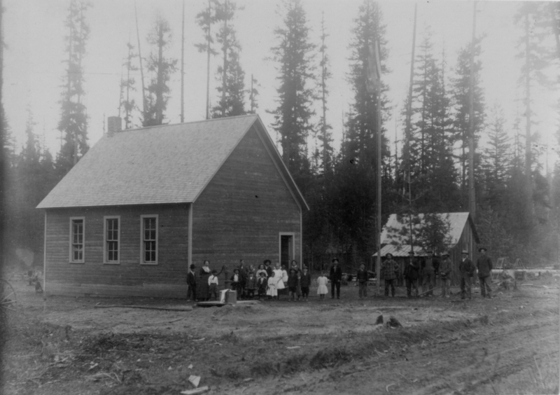 A group of people, including children and adults, stands in front of a small wooden building with three windows and a gabled roof. A smaller structure is visible nearby. Tall trees surround the area, creating a wooded backdrop.
