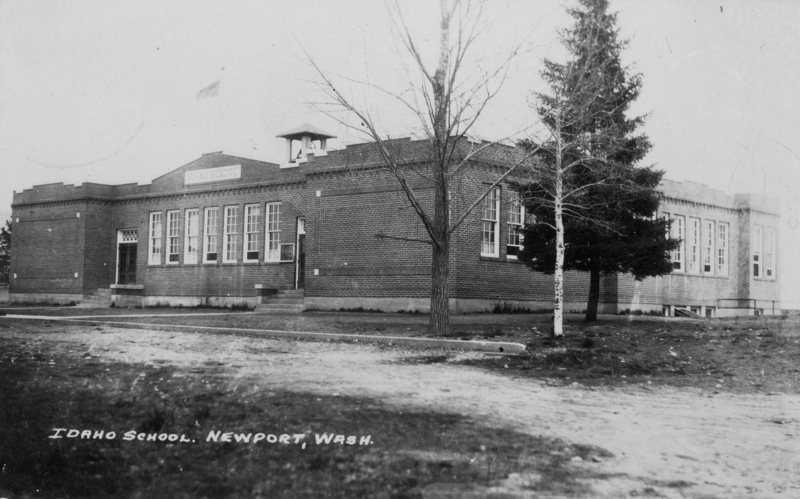 A brick building with large windows and a small bell tower on the roof. There are a few trees in front of the building, and a pathway leading to the entrance. An American flag is flying above the building.  Label near top center of the building reads "IDAHO SCHOOL." Typed text near the bottom left reads "IDAHO SCHOOL. NEWPORT, WASH."