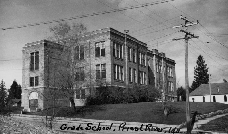 A multi-story brick building with large windows, surrounded by trees and vegetation, set on a grassy hill. A utility pole with power lines is visible in the foreground, and a smaller building with a sloped roof is located nearby. Handwritten text at the bottom reads "Grade School, Priest River, Ida."