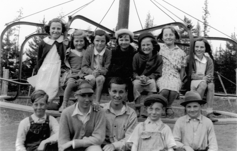 A group of children sit together on a playground structure. There are seven girls in the back row and five boys in the front row. They are wearing a variety of clothing, including dresses, cardigans, hats, and overalls. Trees are visible in the background.