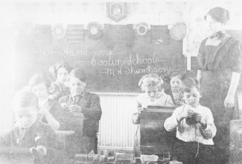 A classroom scene with a group of children sitting at desks, engaged in handwork. A woman stands nearby, possibly a teacher. A chalkboard is in the background with writing that reads "Hand-work" and "Coolin School."