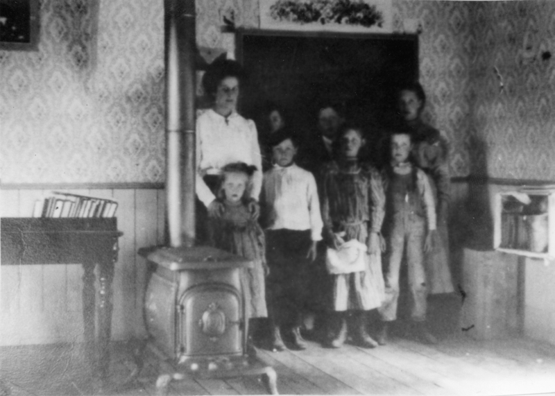 A group of people, including a woman and several children, stand together in a room. There is a wood-burning stove in the foreground, and a small table with books on it to the side. The walls have a patterned wallpaper.
