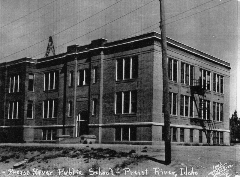 A large brick building with multiple windows and an arched entrance. There is a fire escape on one side. The foreground shows bare ground and a utility pole with wires running above. The building is labeled as "Priest River Public School, Priest River, Idaho."