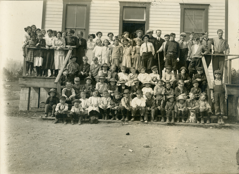 A large group of children and adults gathered on the steps of a building, with some sitting on the ground in front. The people are dressed in early 20th-century clothing, with some wearing hats. The building has visible windows and wooden siding. The scene is outdoors, with dirt ground in the foreground.