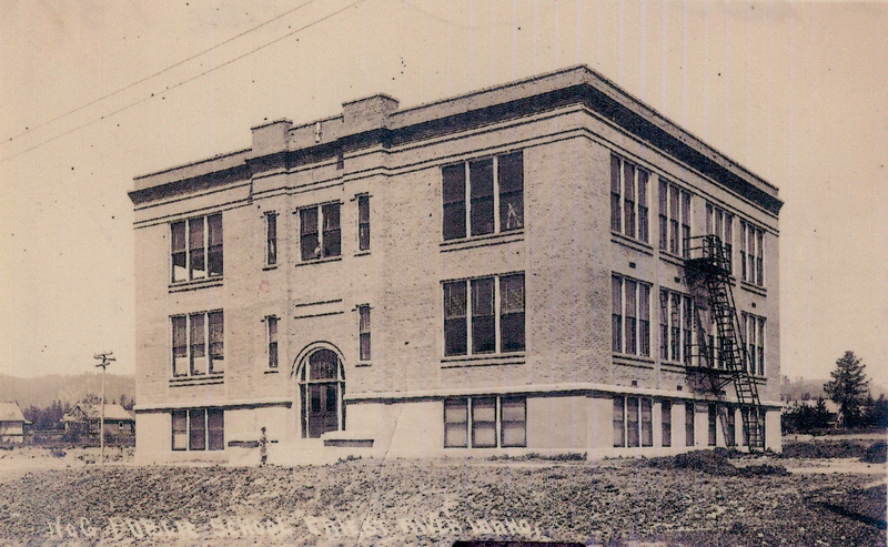 A three-story brick building with large rectangular windows and a fire escape on the side, situated in a grassy area with a few houses and trees in the background.