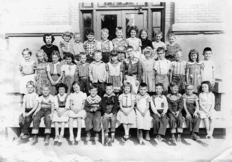 A group of young children arranged in four rows, posing in front of a building with large windows and brick walls. The children are wearing various casual clothing styles typical for school, including dresses, shirts, and sweaters. They are smiling and looking towards the camera.