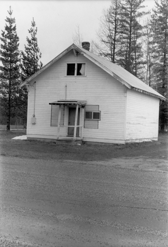 A small, weathered, wooden house with a simple porch stands beside a road. Trees can be seen in the background.