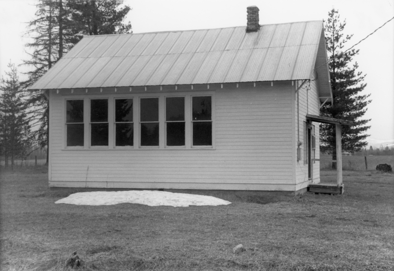 A small building with a metal roof and several windows, situated in a rural area with trees in the background. A patch of snow is on the ground in front of the building.