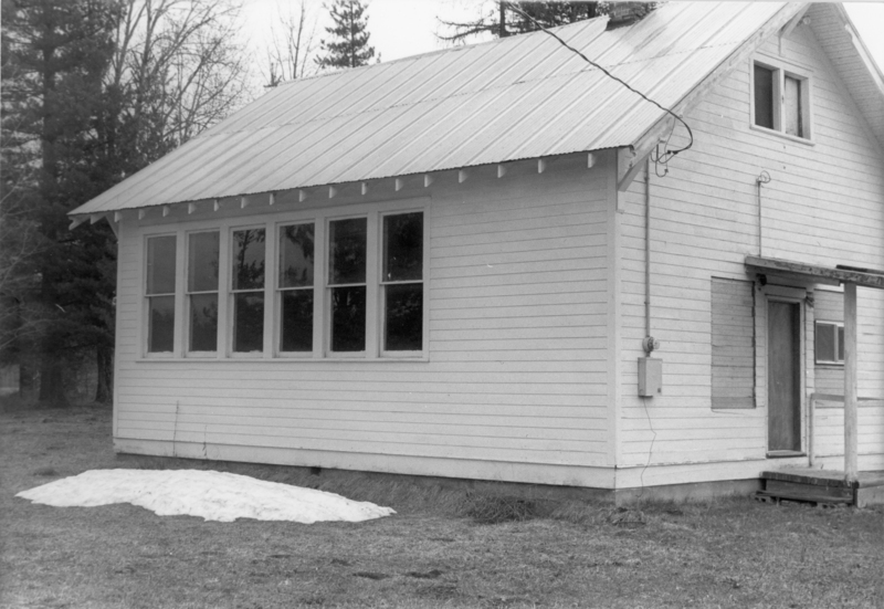 A small wooden house with a metal roof, several windows, and a wooden porch. There is a patch of snow on the ground near the building, and tall trees are visible in the background.