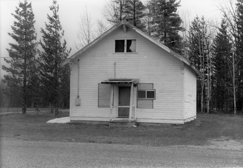 A small wooden house with a front porch, two windows, and a simple door. The house is surrounded by grass and trees, with a forest in the background.