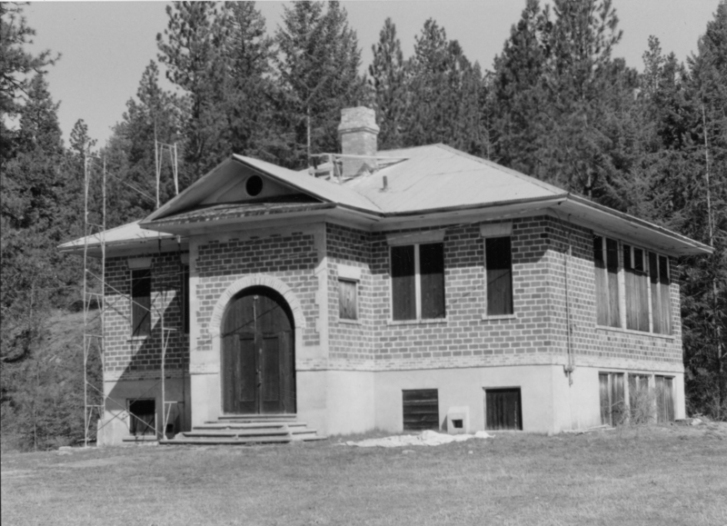 A brick building with a peaked roof and a central chimney, surrounded by trees. The building has an arched wooden door with steps leading up to it and several boarded-up windows.