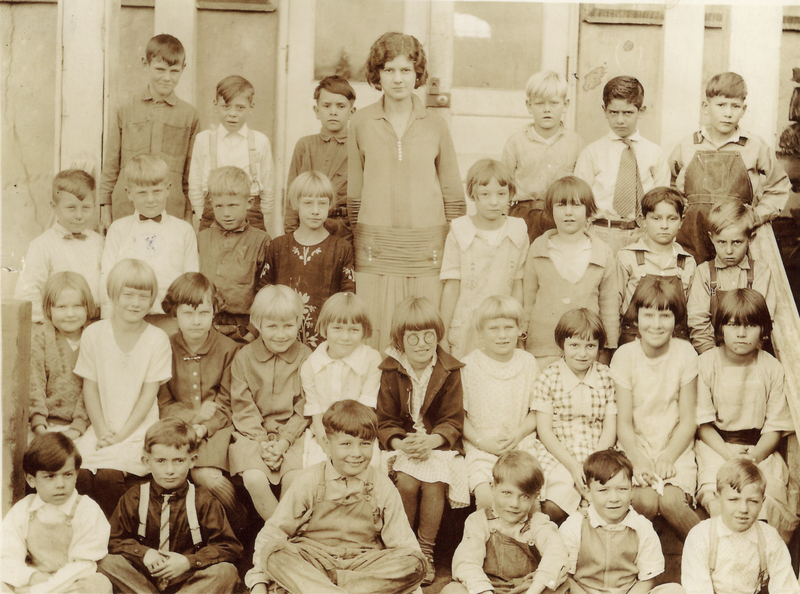 A group of children and one adult, potentially a teacher, are standing and sitting in rows. They are outside in front of a building with a doorway. The children are wearing a variety of clothing styles, including dresses, shirts with ties, and overalls. The adult is standing in the center, surrounded by the children.