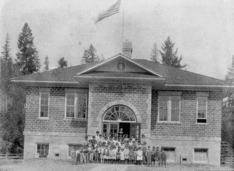A group of children standing in front of a brick building with an arched entrance. Above the building, an American flag is visible. Tall trees are in the background.