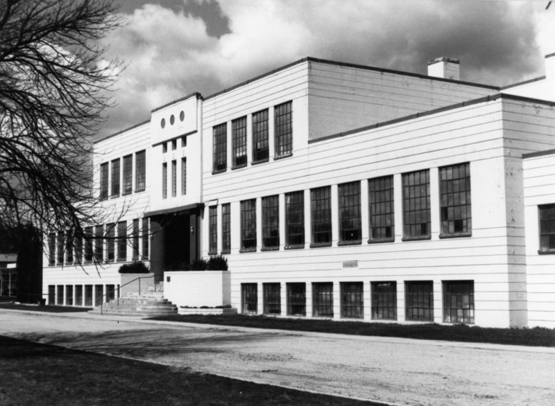 A large two-story building with many windows and a central entrance with steps. There are trees without leaves and a grassy area nearby. The sky has some clouds.