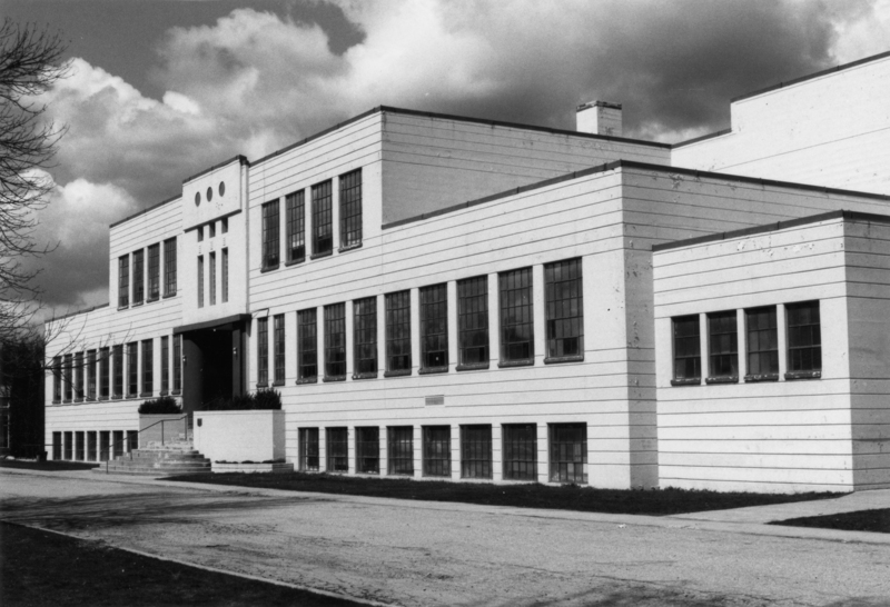 A large, rectangular building with many windows and a flat roof. The structure is two stories high with an entrance featuring steps and a few decorative elements above it. A paved path leads to the entrance, and there are small plants or shrubs near the doorway. The sky has some clouds.