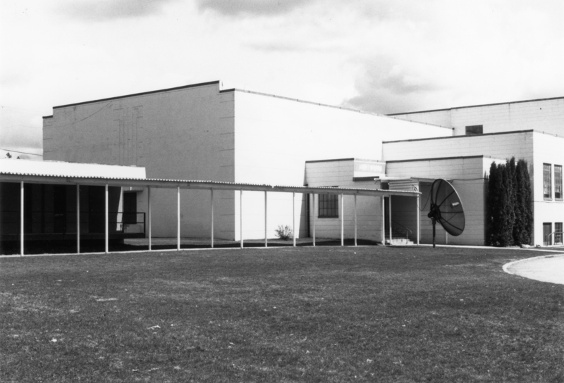 A large building with a flat roof and a long covered walkway along the front. A large satellite dish is mounted next to the building. The surrounding area is a grassy lawn.