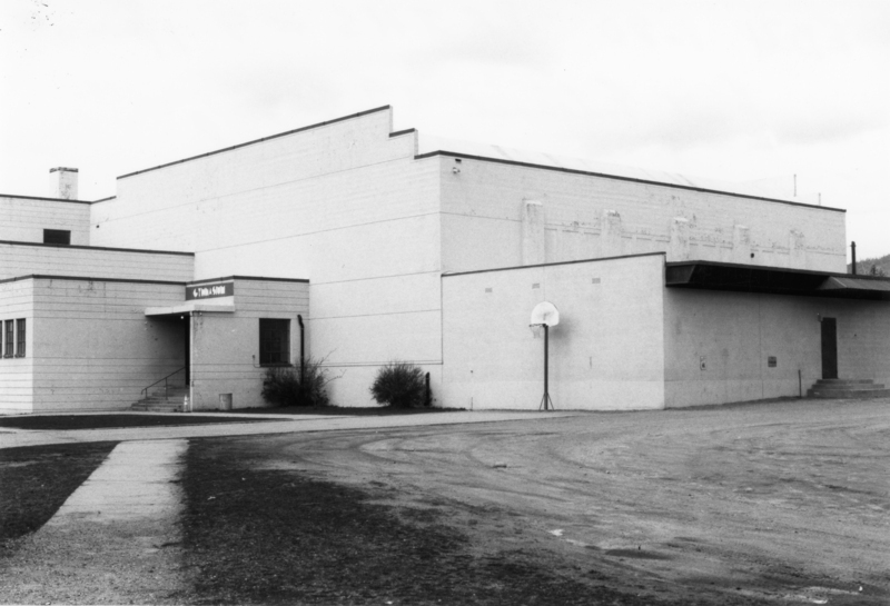 A large rectangular building with a small entrance canopy on the left and a basketball hoop mounted on the wall to the right. The ground is rough and the area is bordered by some shrubs. Sign above entrance reads: "Gymnasium".